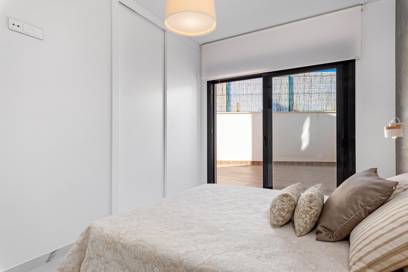 Bright modern bedroom with white wardrobes, a beige patterned bedspread, and decorative pillows by a large sliding glass door to a balcony outside the room.