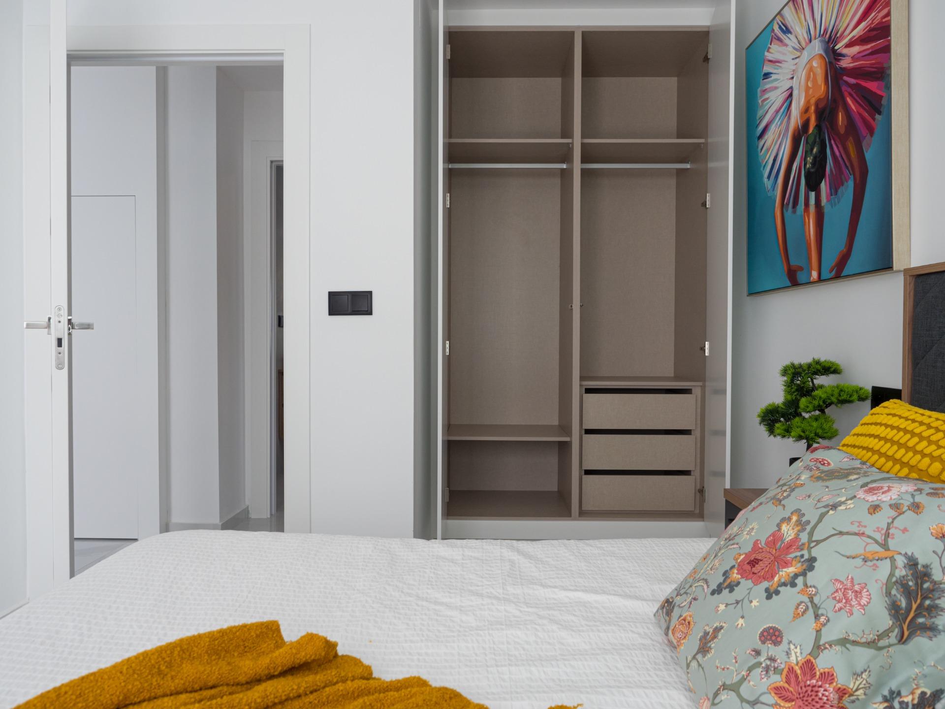 Bedroom with an open beige wardrobe showing shelves and drawers, floral bedding, and a bright colorful painting on the wall to the right.