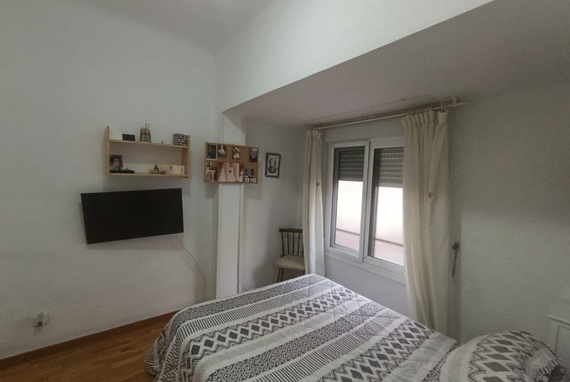 Bedroom with two twin beds in geometric-pattern quilts, a wall-mounted flat-screen TV, and a window with cream curtains nearby.
