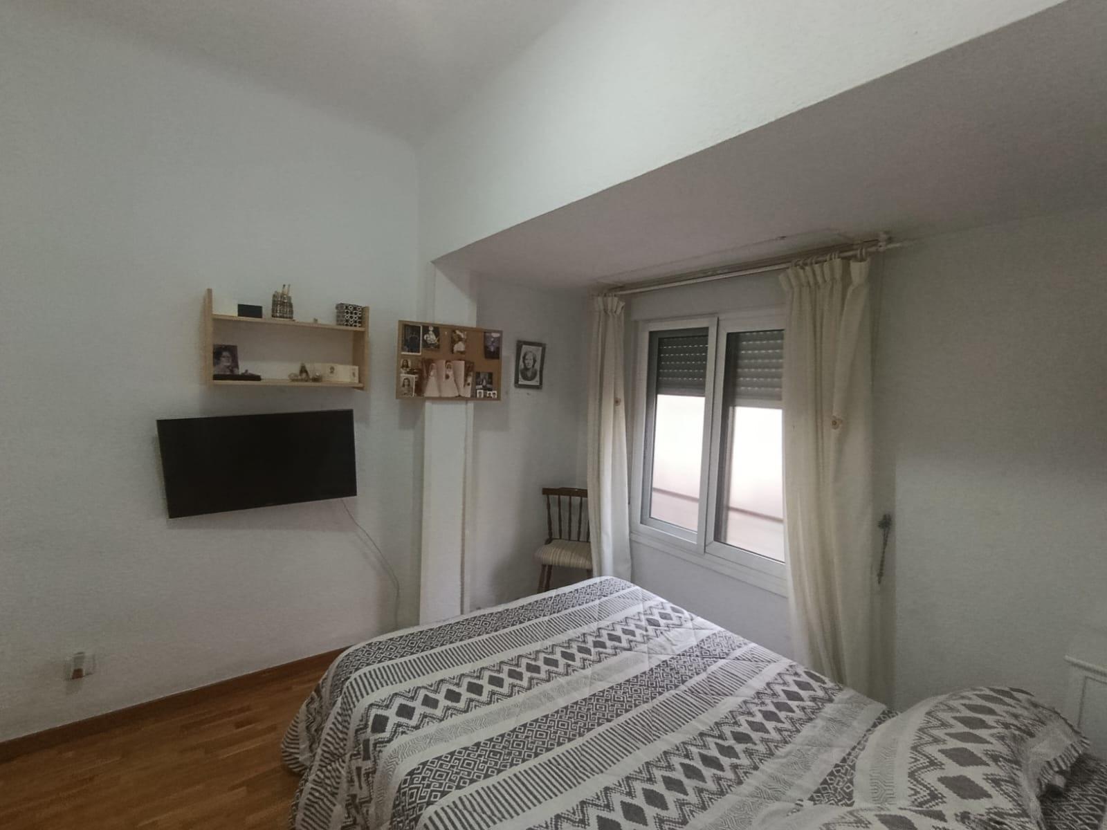 Bedroom with two twin beds in geometric-pattern quilts, a wall-mounted flat-screen TV, and a window with cream curtains nearby.