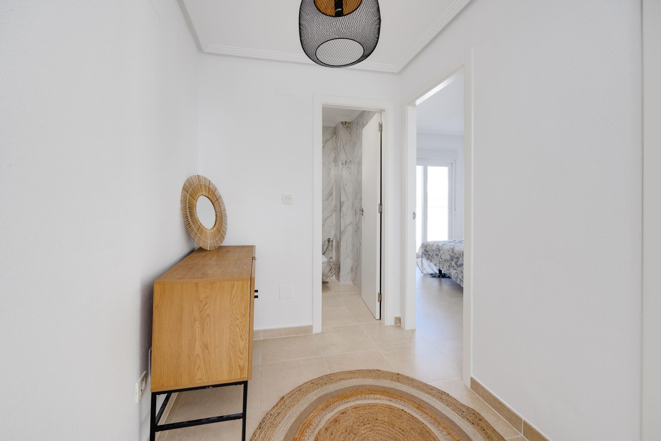 White entry hallway with a wooden console and circular braided mirror, leading to a marble-tiled bathroom and a sunlit bedroom beyond.