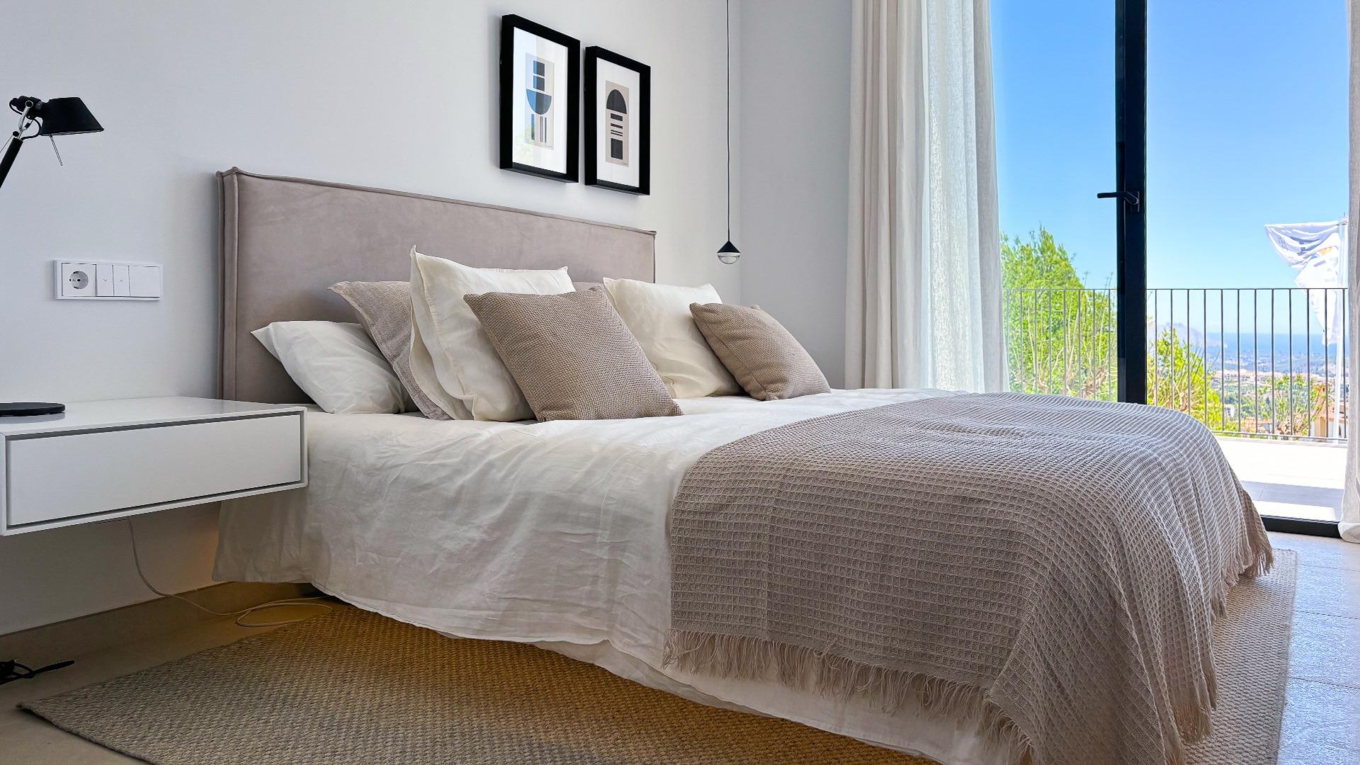 Bright hotel-style bedroom with white bedding, beige throw, and a balcony view of mountains and blue sky beyond the door.