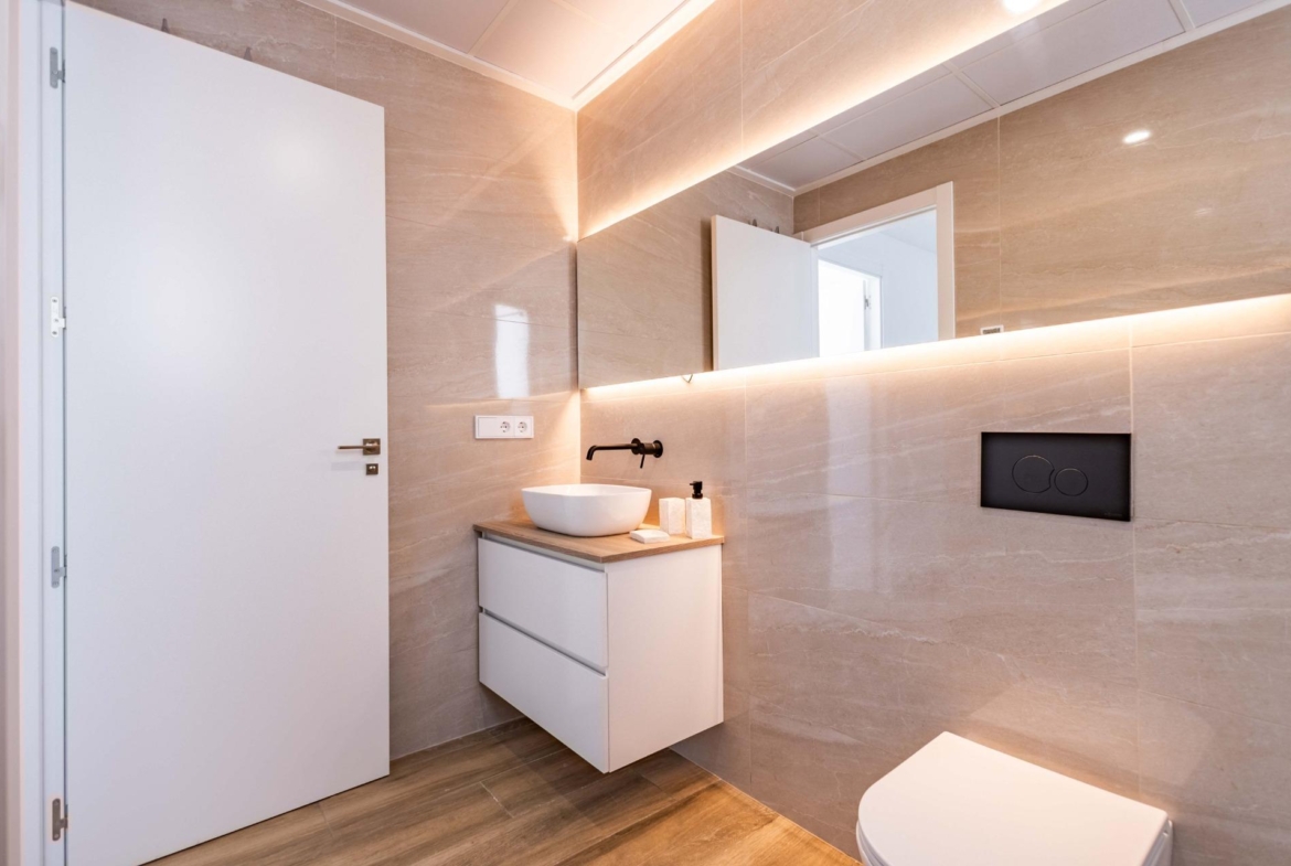 Modern beige-tiled bathroom with a white vanity, vessel sink, and wall-mounted faucet under a backlit mirror shelf
