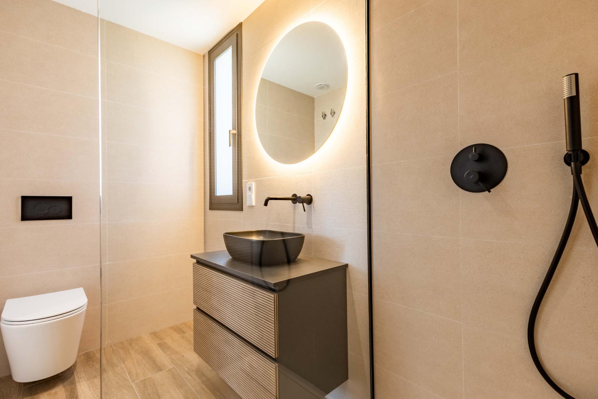 Modern beige-tiled bathroom with a round backlit mirror above a dark vanity and black vessel sink; wall-mounted faucet visible on the sink curbside feature.