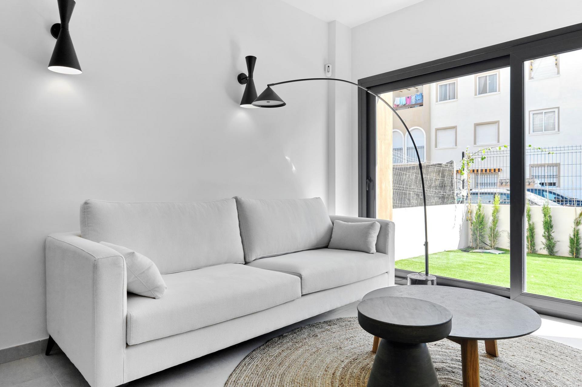 Modern living room with a light gray sofa, black arc floor lamp, and a large glass window showing a green yard outside at the patio