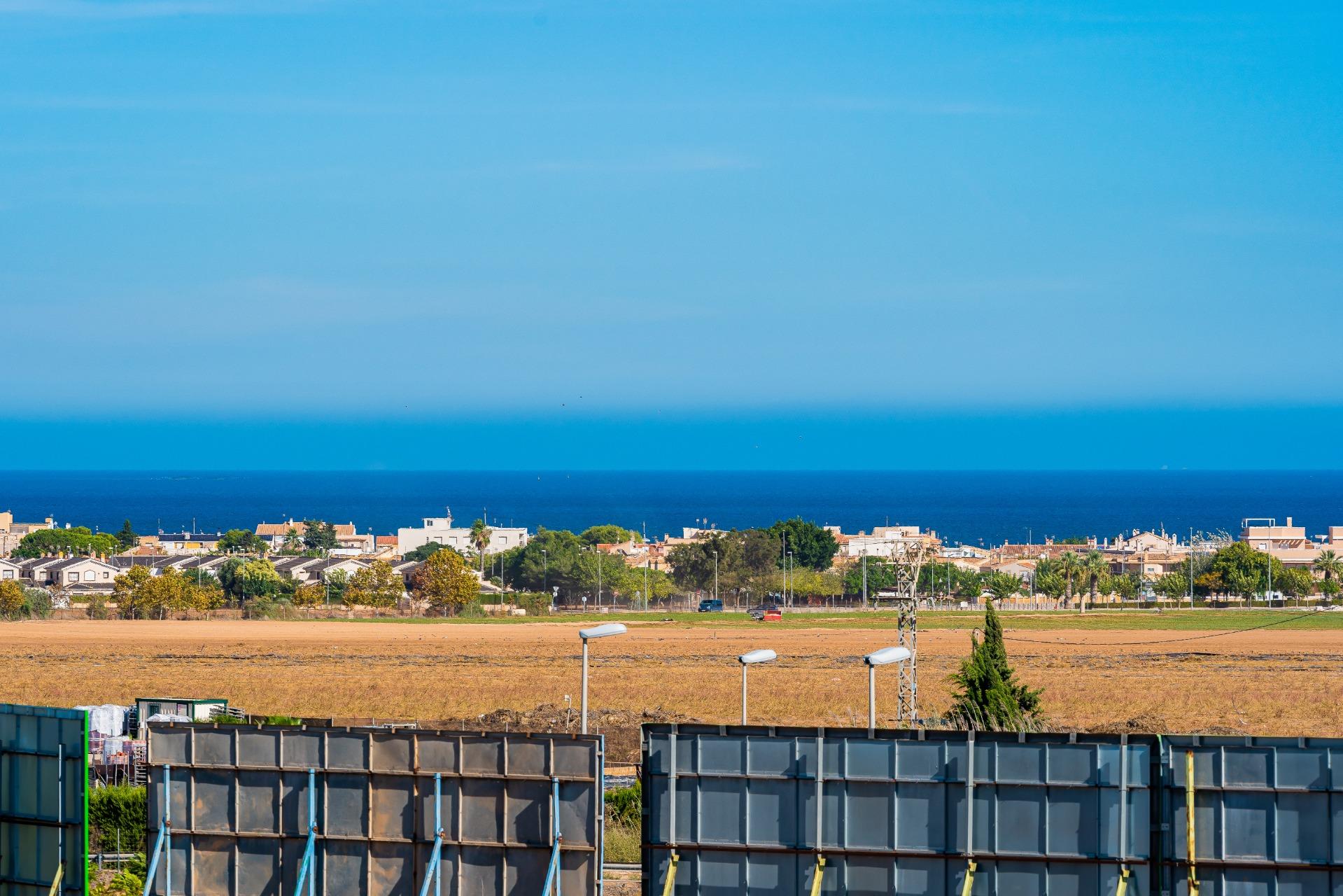 Industrial fence panels and streetlights in the foreground with a coastal town and blue sea beyond under a clear sky.]"