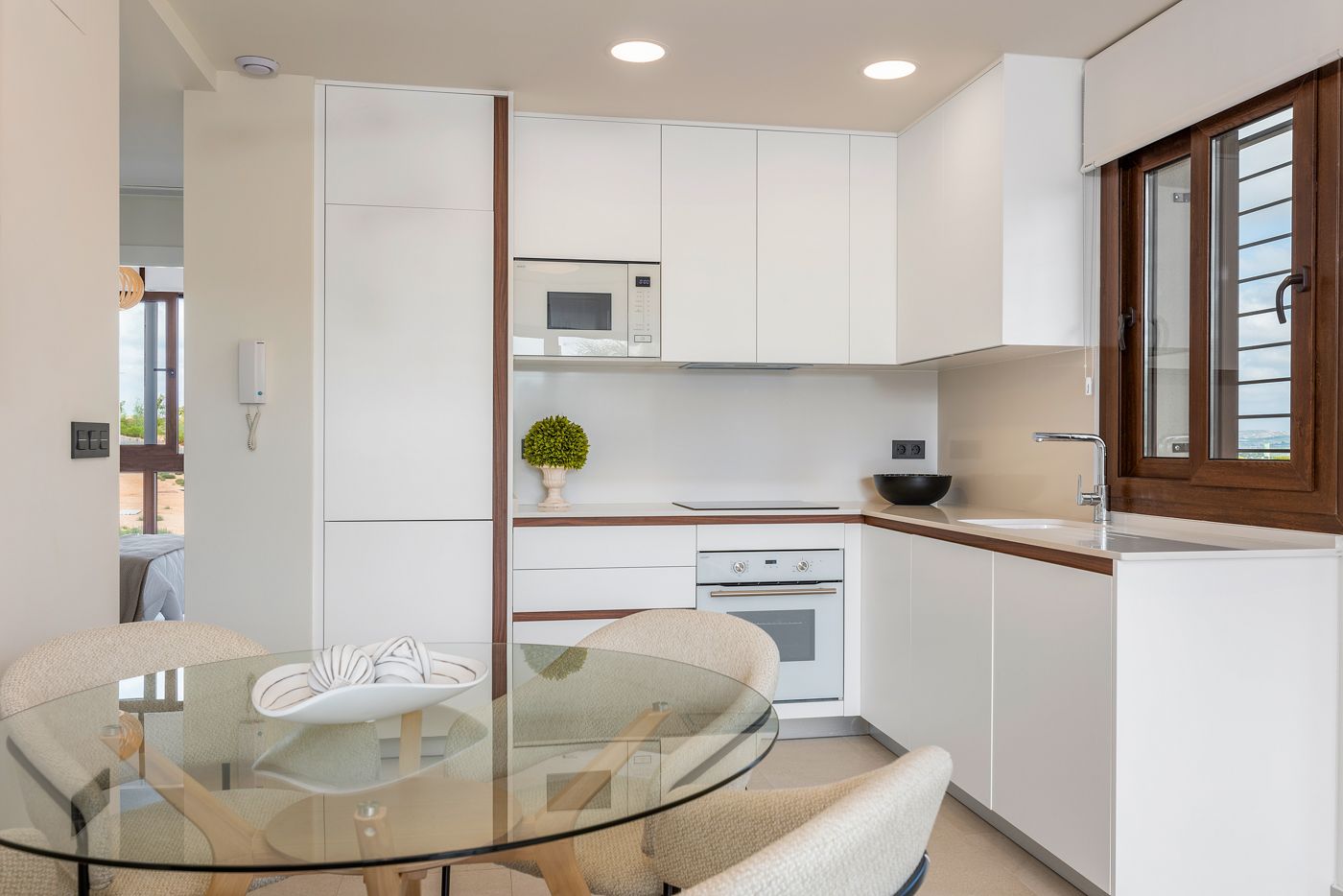 Modern white kitchen with wooden trim, built-in microwave and oven, and a glass round dining table in the foreground.