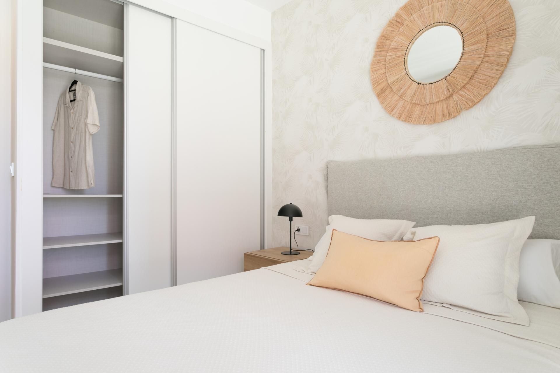 Neutral bedroom with a white sliding-door wardrobe, a beige shirt hanging inside, and a round woven straw mirror above the bed.