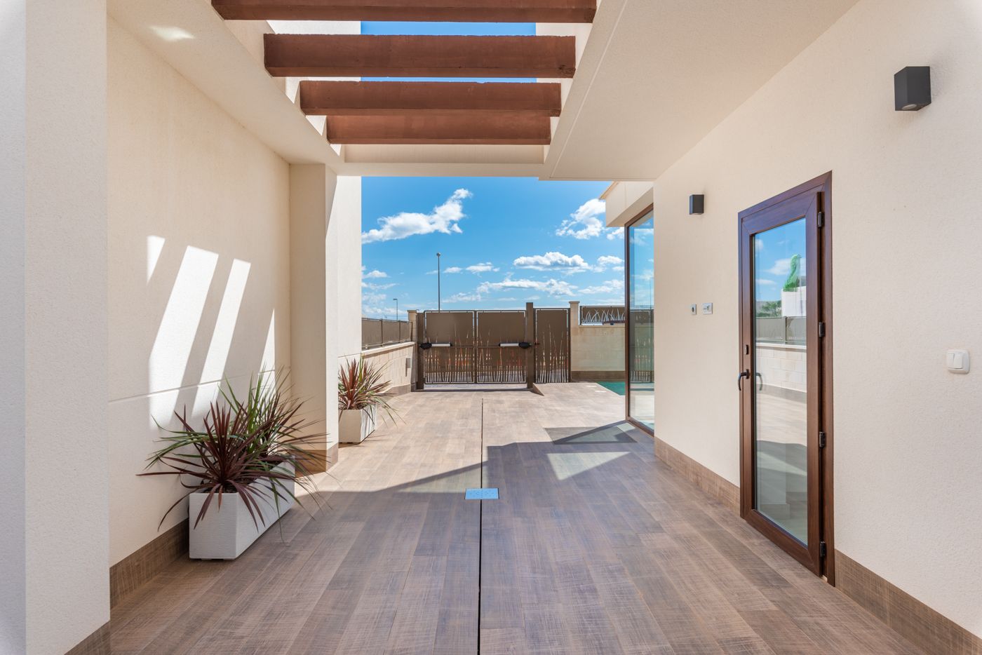 Open-air patio corridor with wooden-beam pergola, beige walls, tiled floor, and planters along the sides leading to a gate. not after.