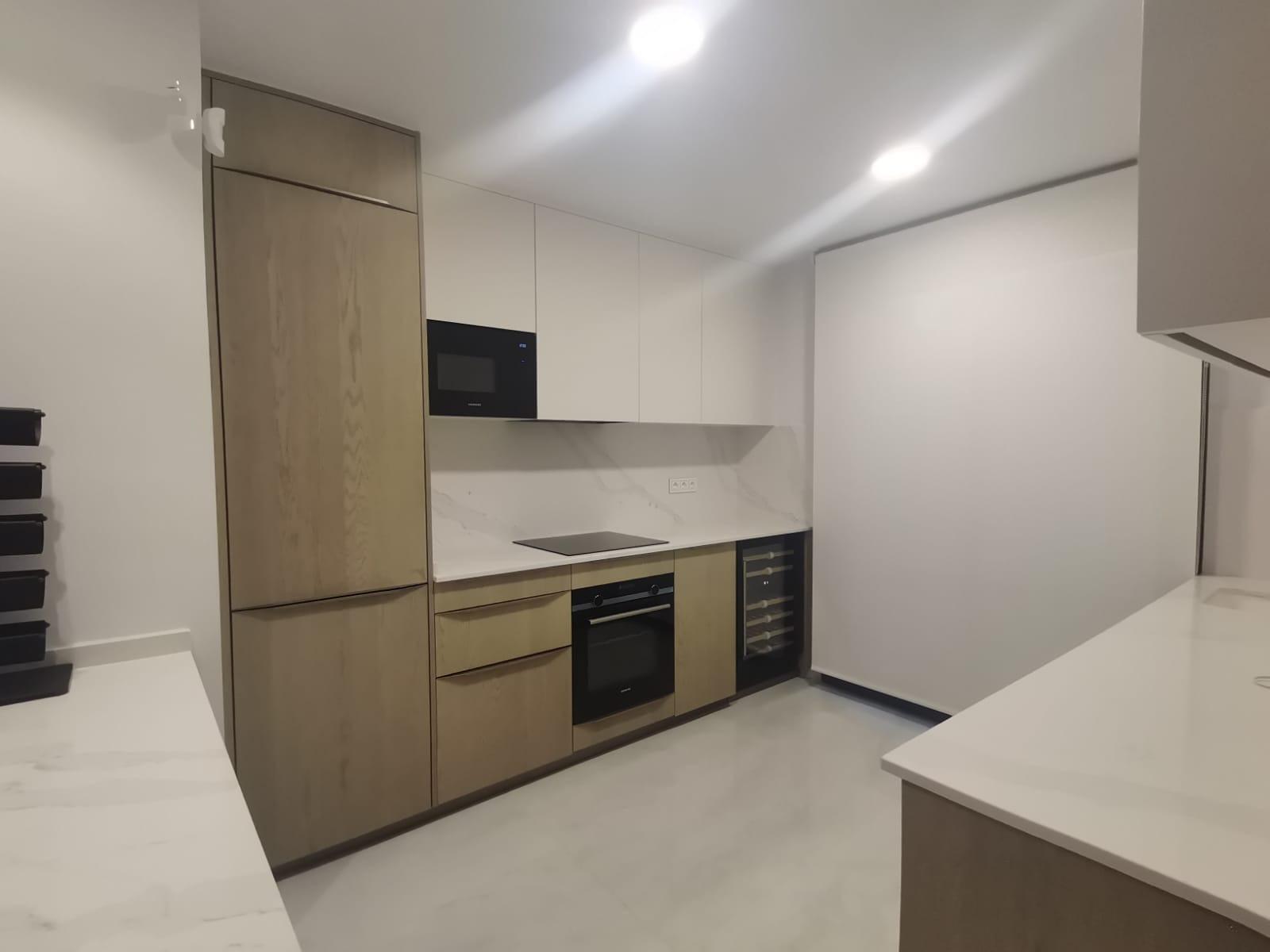 Modern minimalist kitchen with white upper cabinets, wood-toned tall cabinet, integrated microwave and oven, induction hob, marble backsplash, and a wine cooler at the end