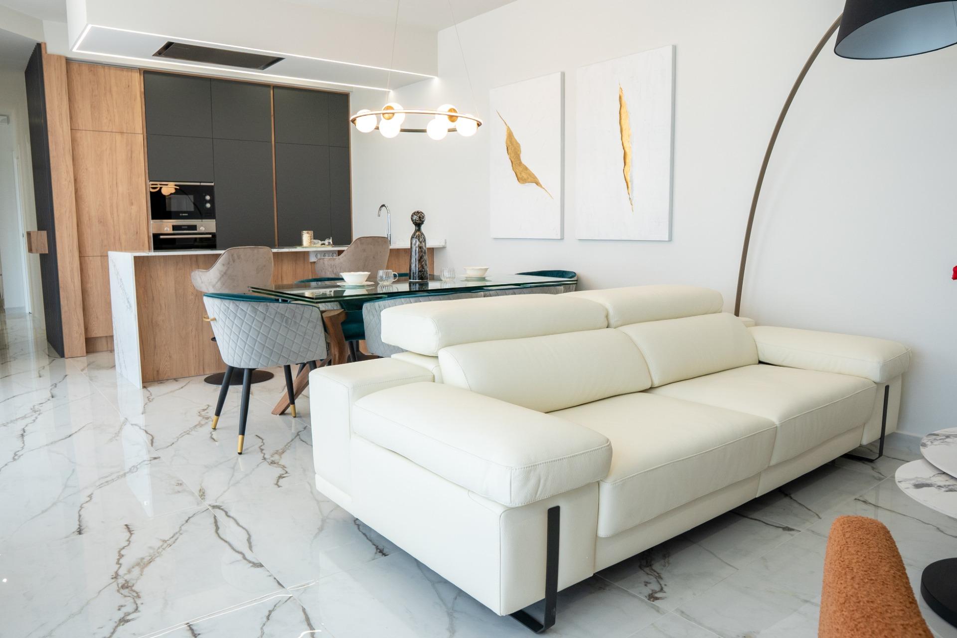 Open-plan living area with a white leather sofa, glass dining table, and wooden kitchen cabinets in the background.