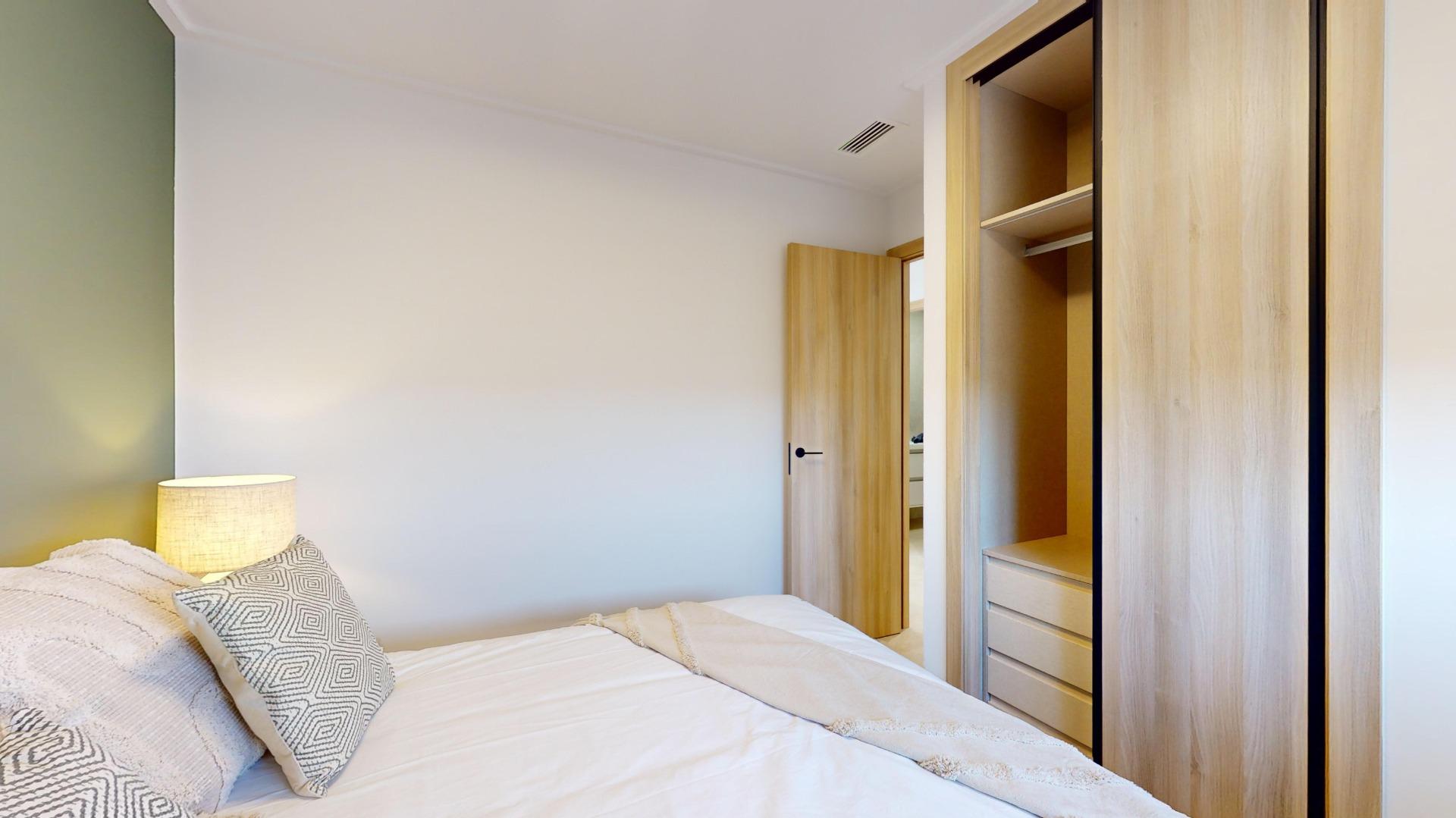 Bedroom with a bed and patterned pillows, a glowing bedside lamp on the left, and an open wooden wardrobe on the right with shelves and drawers.