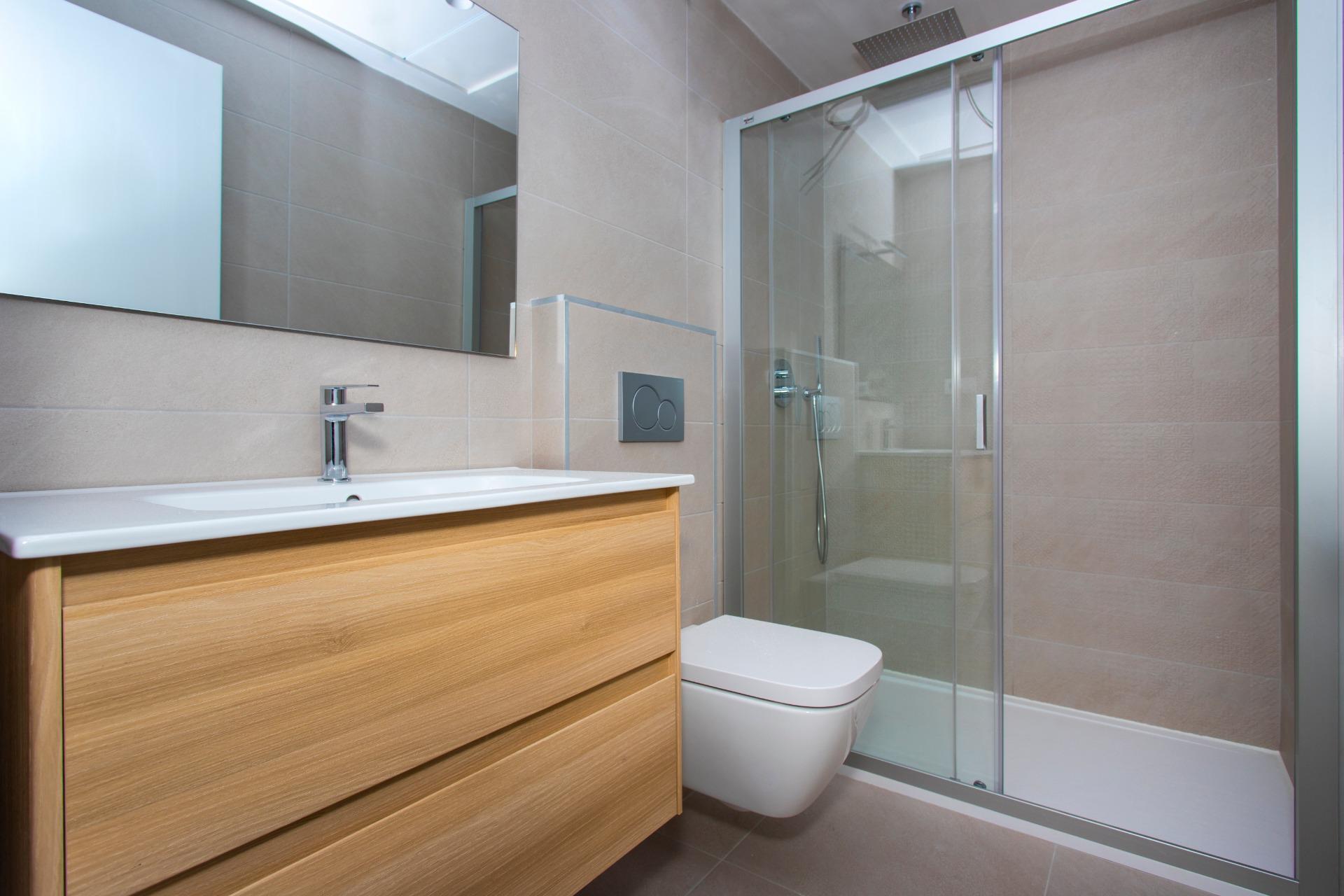 Modern bathroom with a wooden vanity, white sink, wall-mounted toilet, and glass shower enclosure.