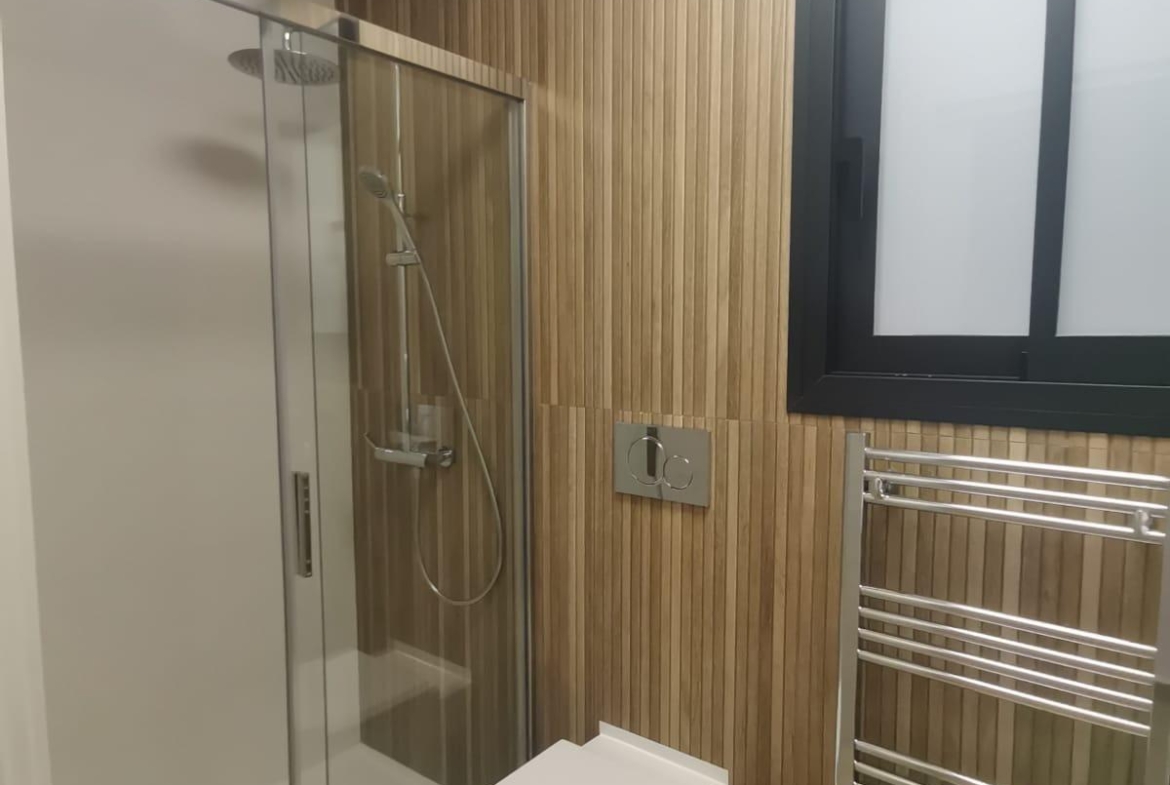 Modern bathroom with a glass shower enclosure, vertical wood-paneled wall, wall-mounted dual-flush toilet, and chrome towel warmer under a frosted window