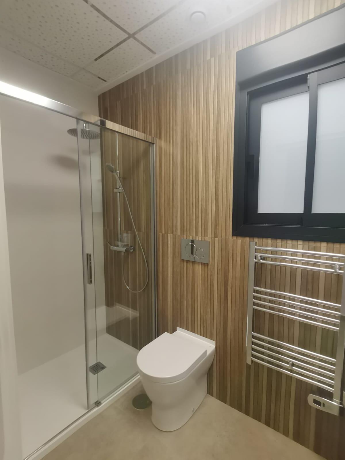 Modern bathroom with a glass shower enclosure, vertical wood-paneled wall, wall-mounted dual-flush toilet, and chrome towel warmer under a frosted window