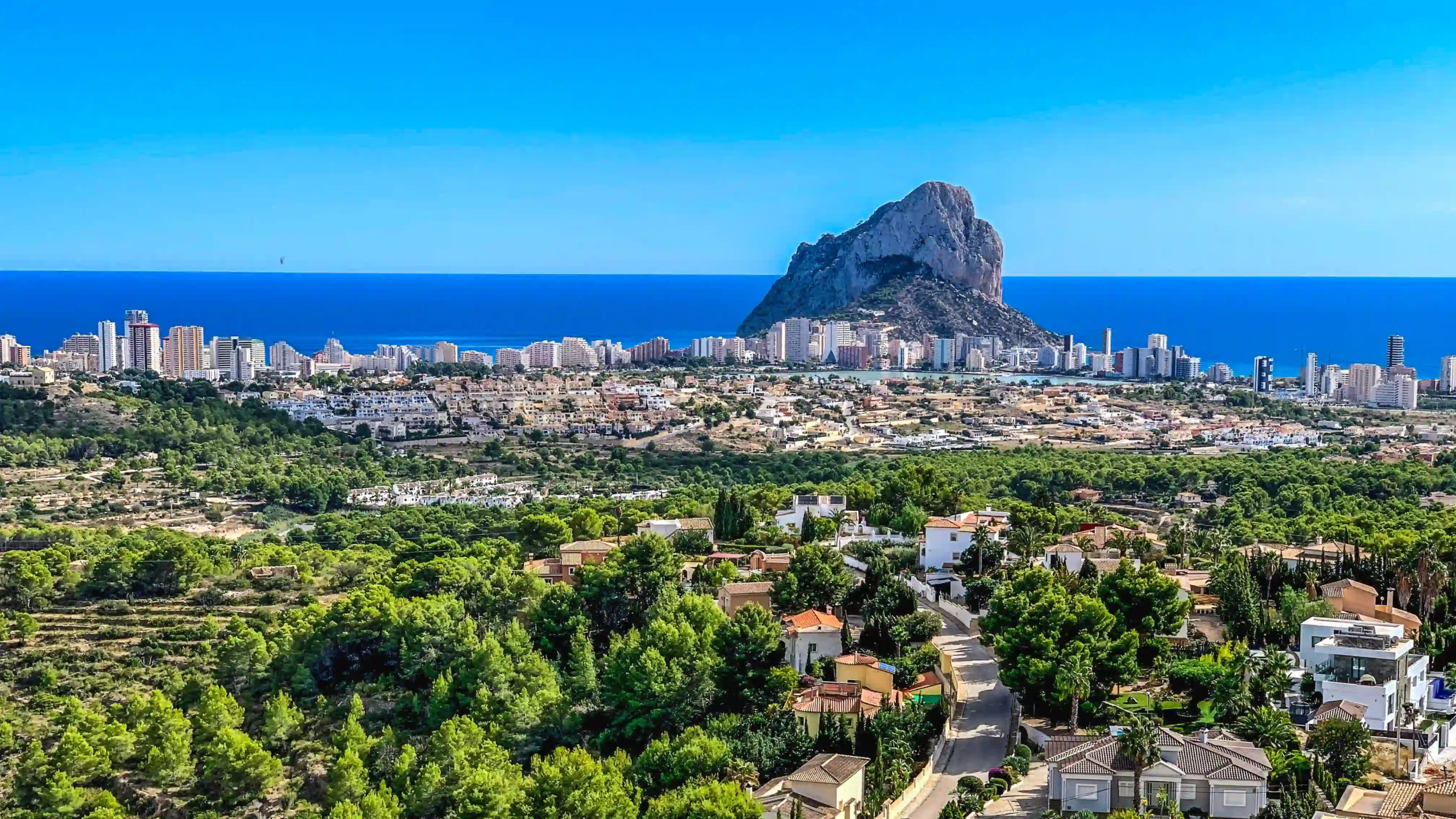 Coastal city skyline with high-rise buildings along the blue sea and a large rocky rock jutting from the shoreline.