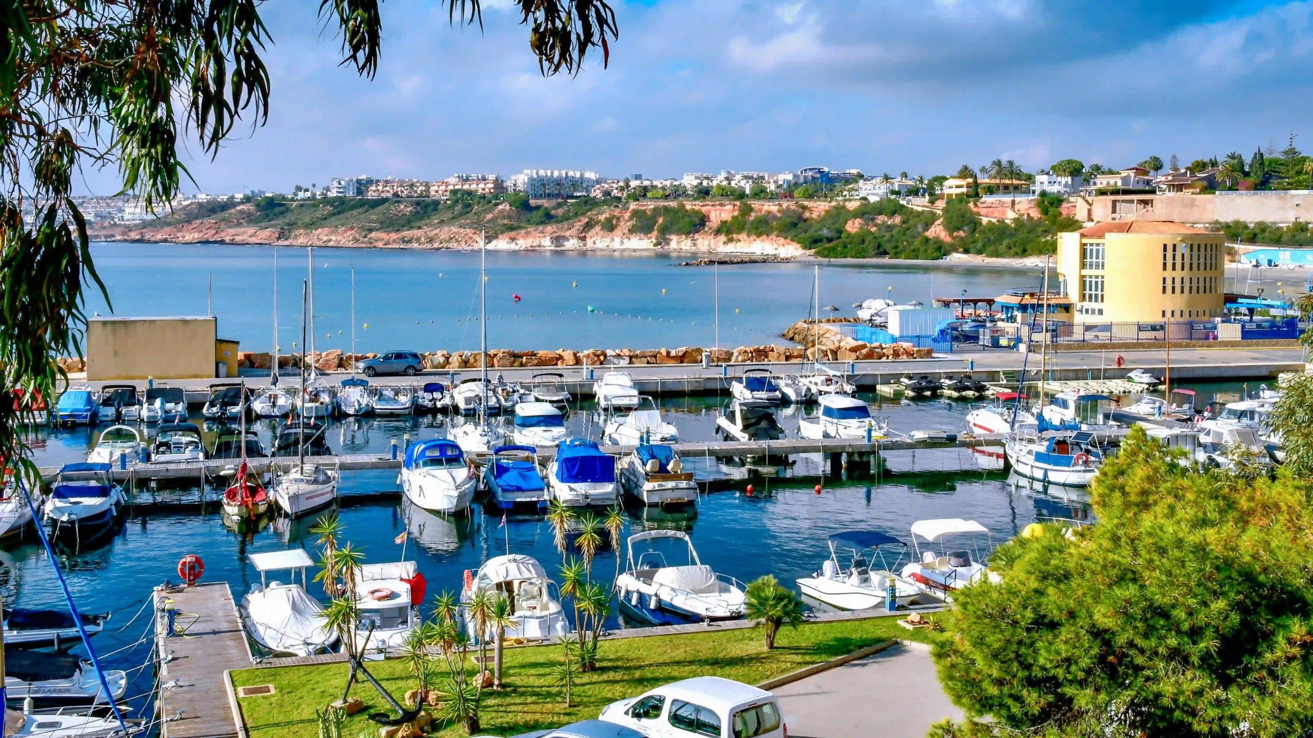 Marina filled with sailboats and yachts, calm blue water, rocky shoreline, and a hillside town in the distance.