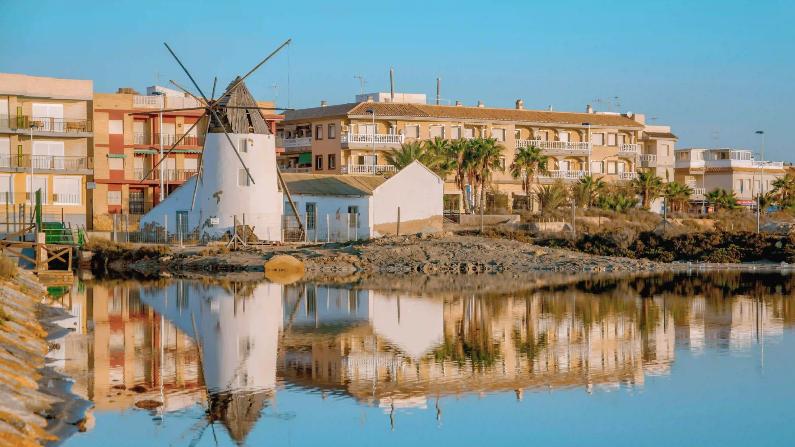 Waterfront townscape with a white windmill-like structure, pastel buildings, and palm trees, all reflected in calm harbor water under a blue sky.