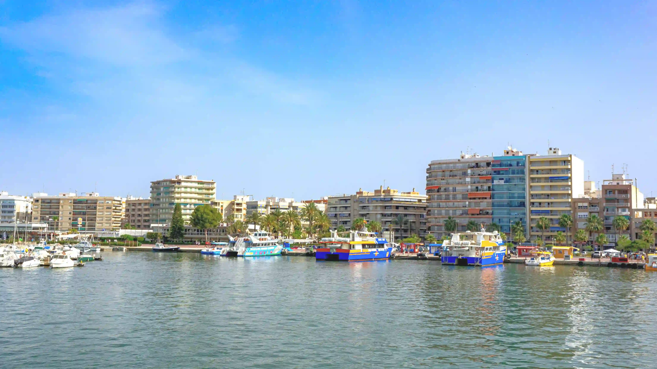 Harbor scene with boats docked along a city waterfront and mid-rise buildings in the background under a clear blue sky.