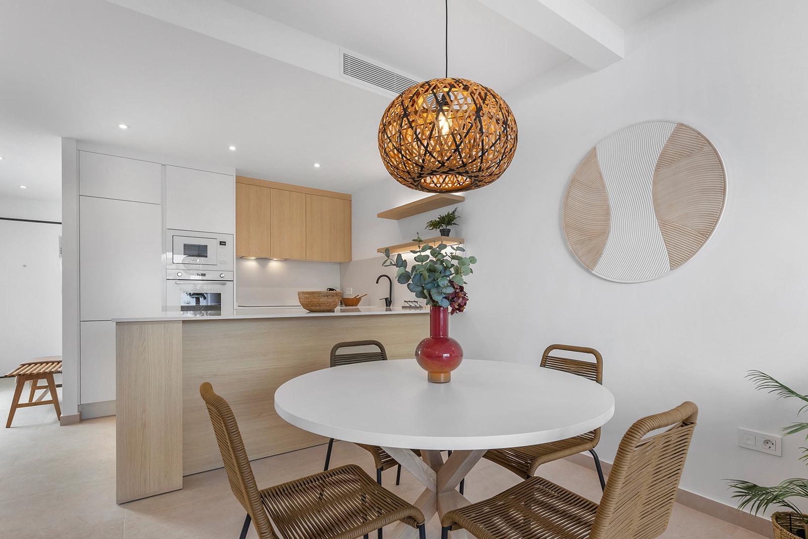 Open-plan kitchen and dining area with white cabinets, wooden island, round white table, and wicker chairs under a woven pendant light.
