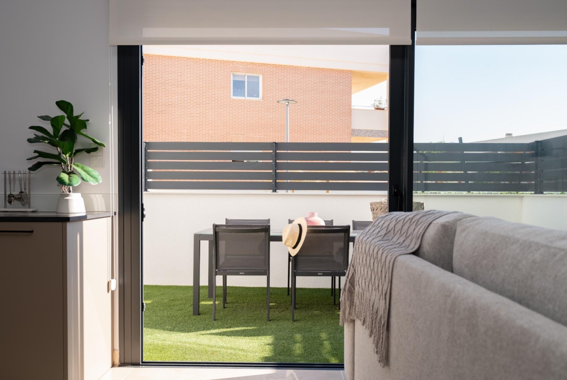 Indoor living space with a large sliding glass door opening to a small patio, showing a table with four chairs on artificial grass and a hat resting on one chair; potted plant on the counter to the left.