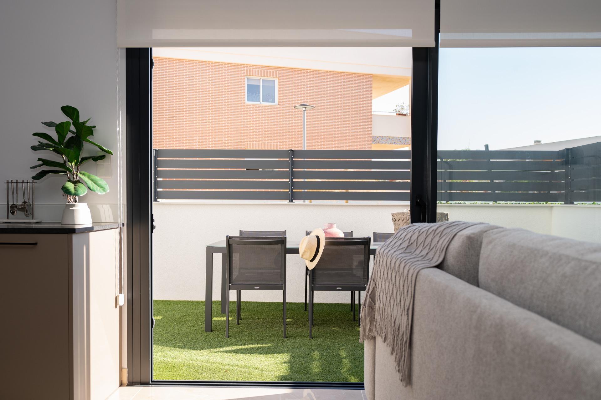 Indoor living space with a large sliding glass door opening to a small patio, showing a table with four chairs on artificial grass and a hat resting on one chair; potted plant on the counter to the left.