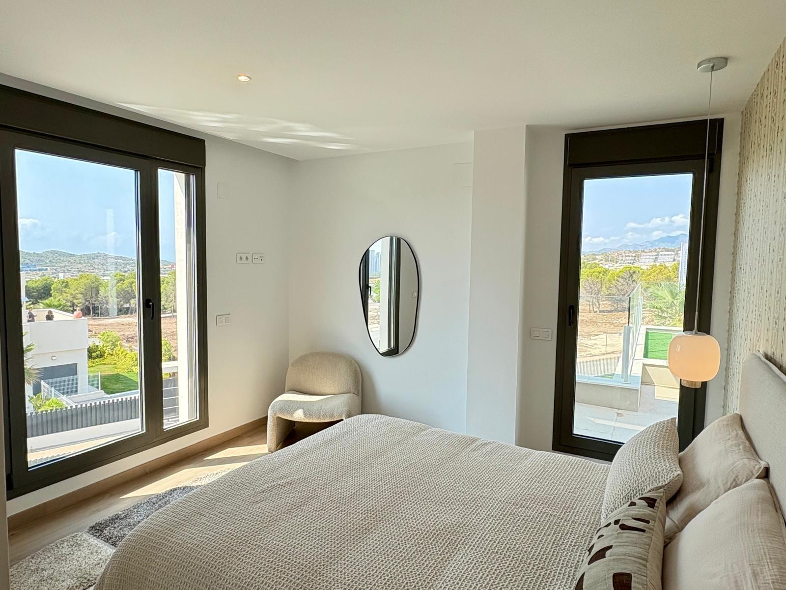 Bright modern bedroom with large framed windows showing outdoor landscape; bed in foreground and a round wall mirror.