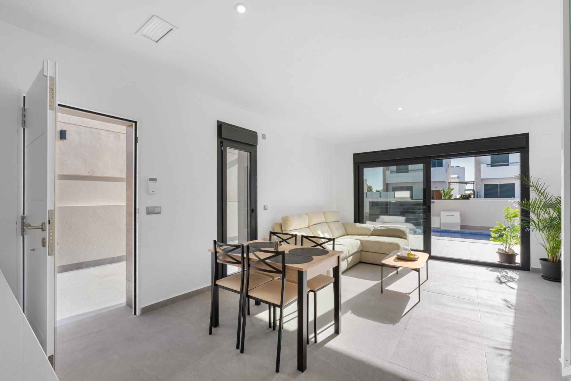 Bright, minimalist living and dining area with a beige sofa, black-framed sliding doors, and a view of the pool outside.