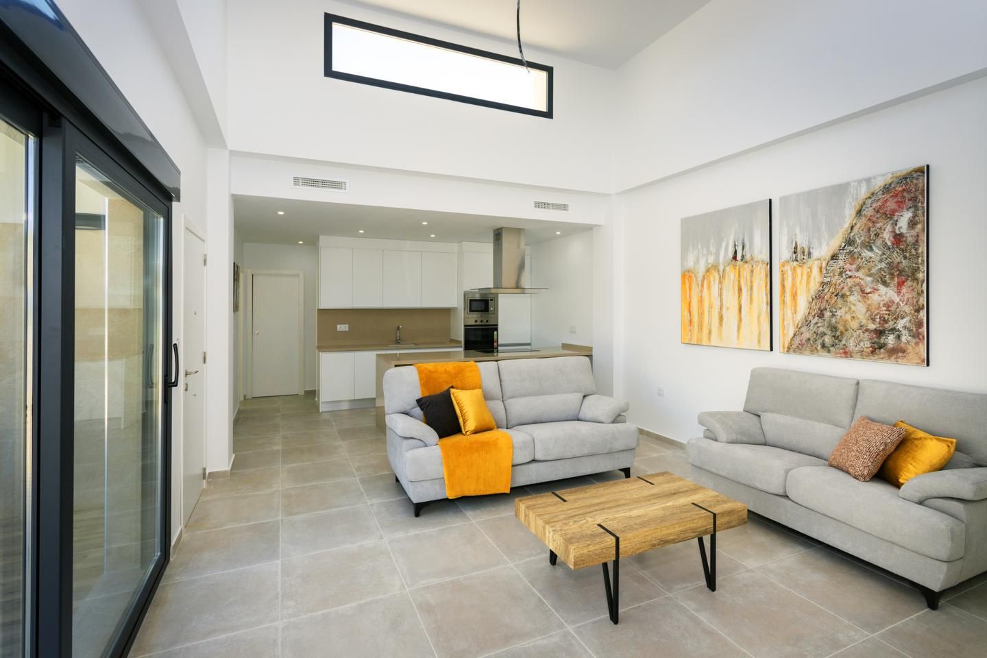 Open-plan living room with two light gray sofas, orange throw blankets and cushions, a wooden coffee table, and abstract wall art; white kitchen in the background and large glass doors to the left.