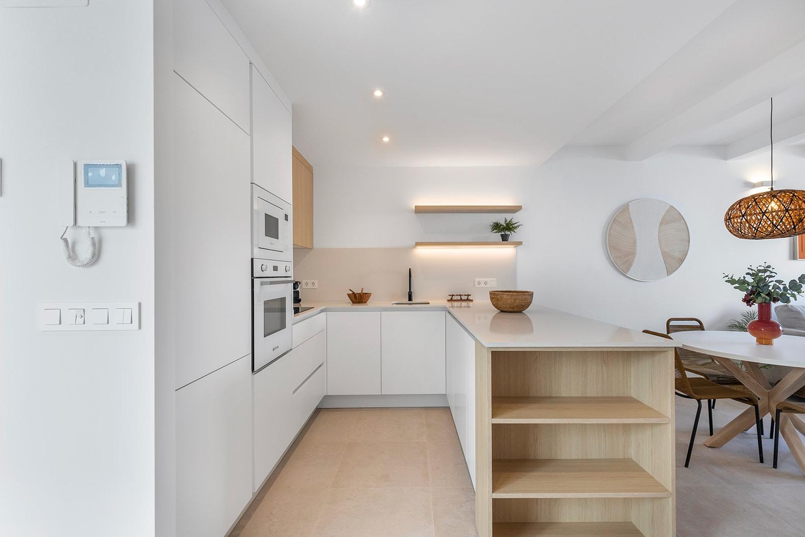Bright white, open-plan kitchen with built-in ovens, pale wood island, and a surrounding dining area with a wicker pendant light.