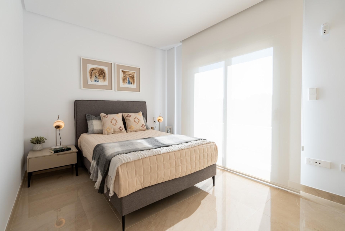 Bright minimalist bedroom with gray upholstered bed, beige quilt, patterned pillows, and two framed artworks above the headboard.