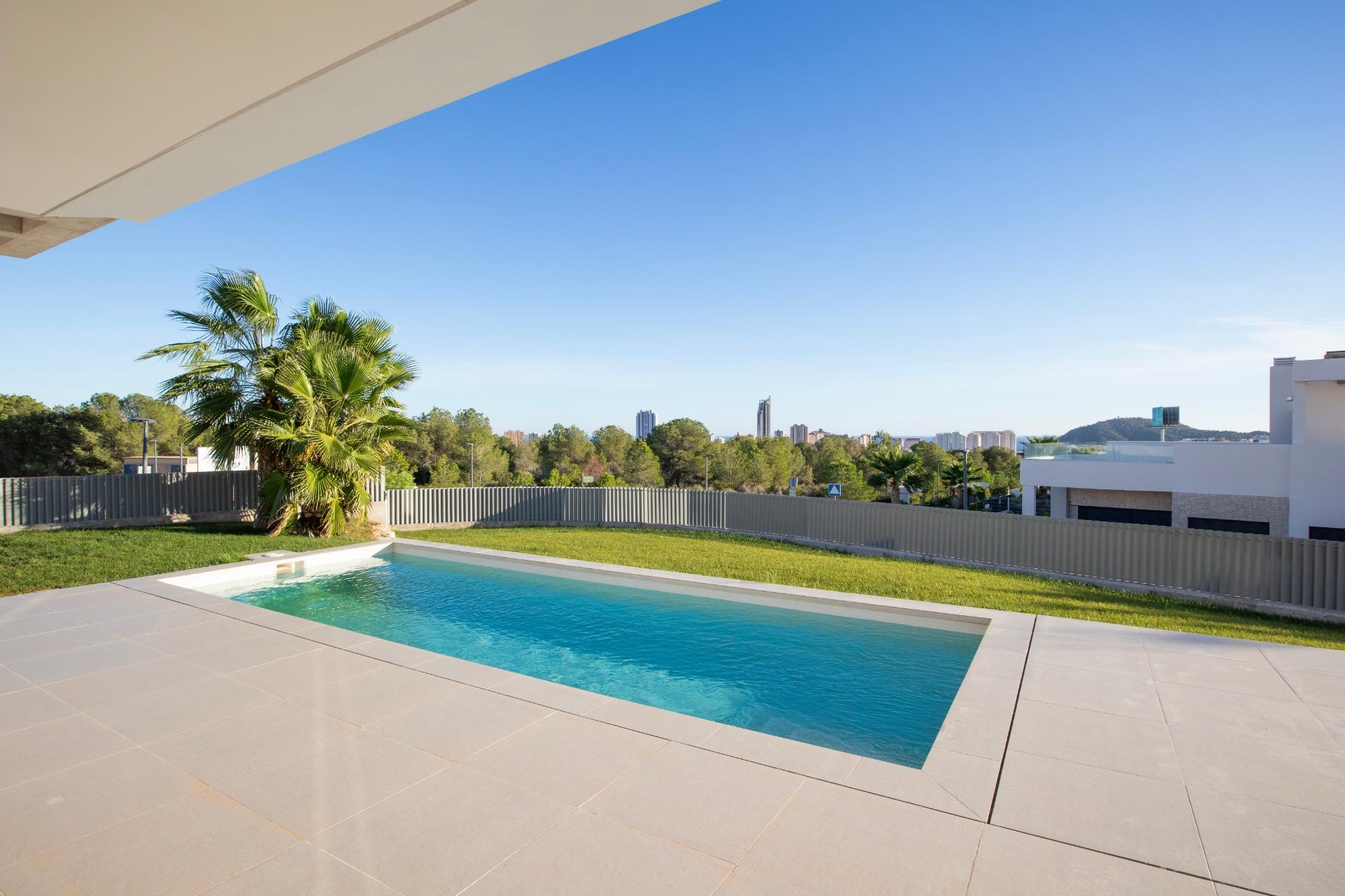 Modern rectangular pool surrounded by beige stone tiles, a palm tree, and a white fence with a distant city skyline under a clear blue sky.