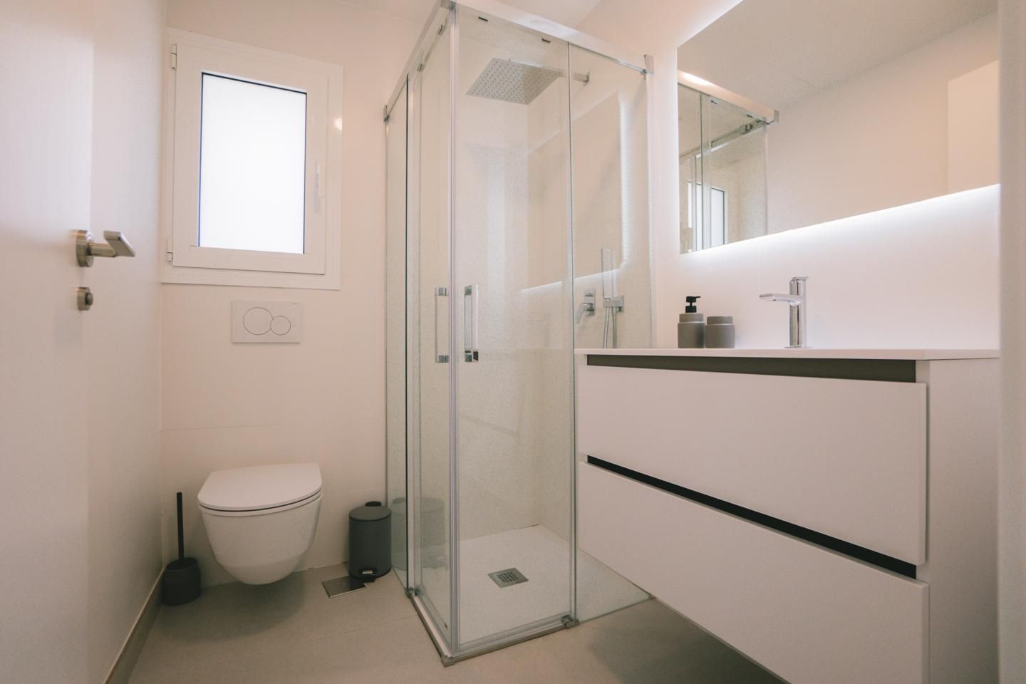 Minimalist white bathroom with a corner glass shower, wall-hung toilet, and floating vanity with a backlit mirror.