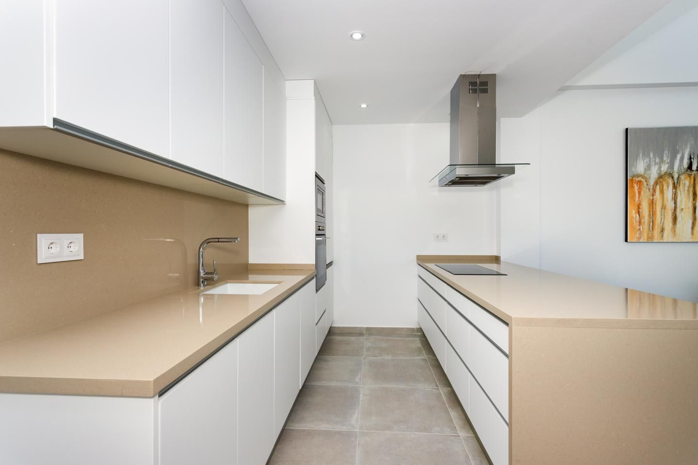 Modern galley kitchen with beige countertops, white cabinets, and a central island; stainless steel range hood overhead.