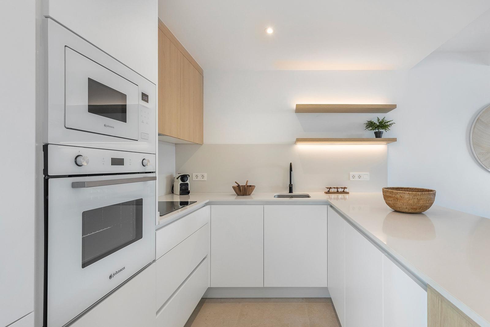 Modern white kitchen with built-in double ovens, light wood upper cabinet, and an L-shaped countertop.