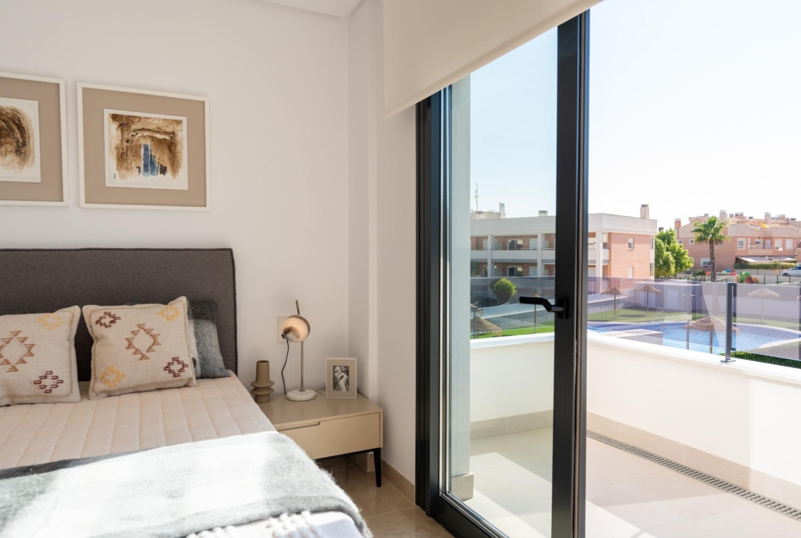 Bright bedroom with a bed, nightstand, and two framed abstract artworks; a large glass sliding door opens to a sunny balcony with a pool view.