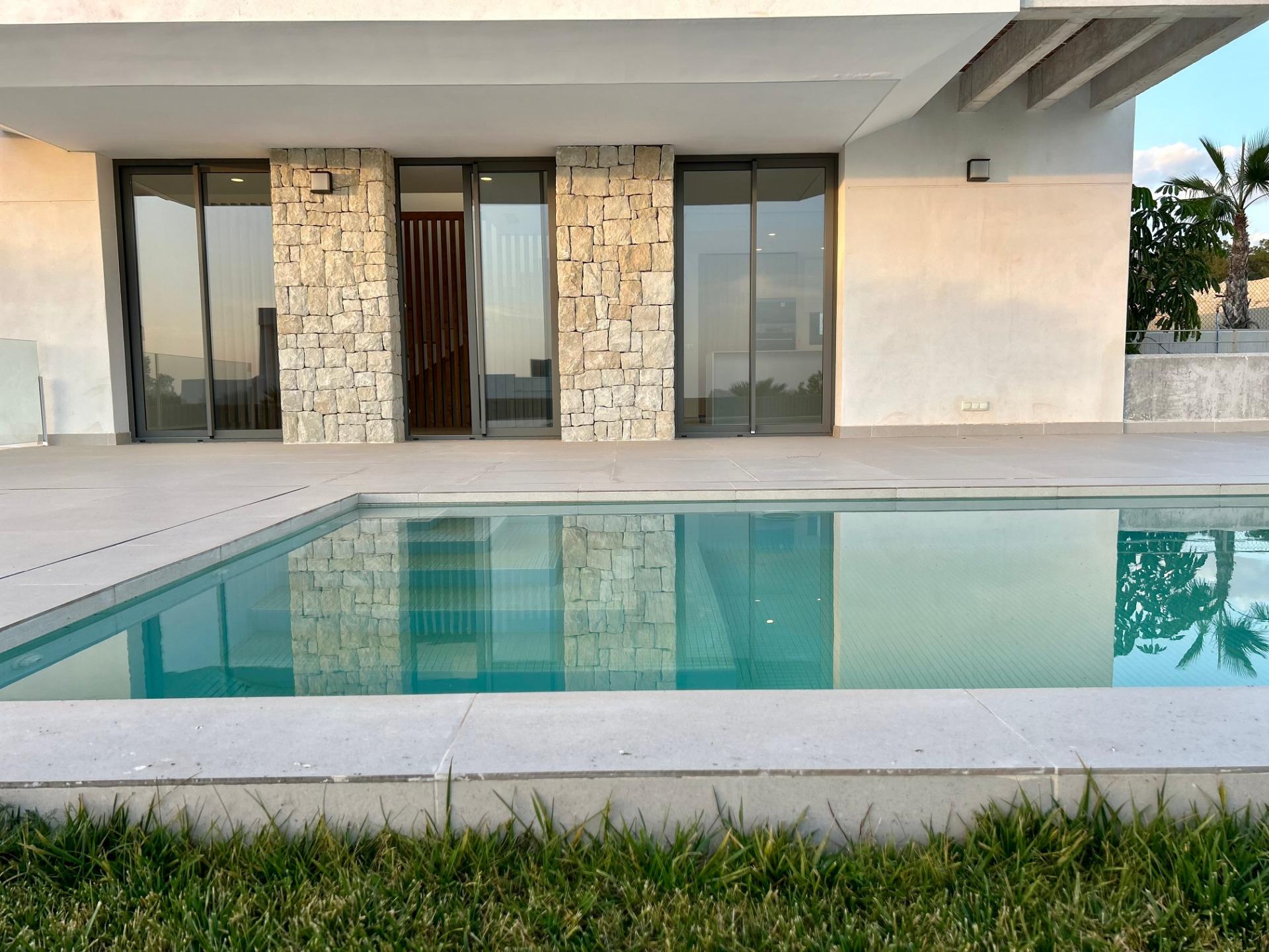 Modern house frontage with large glass sliding doors, stone columns, and a rectangular pool in the foreground reflecting the building