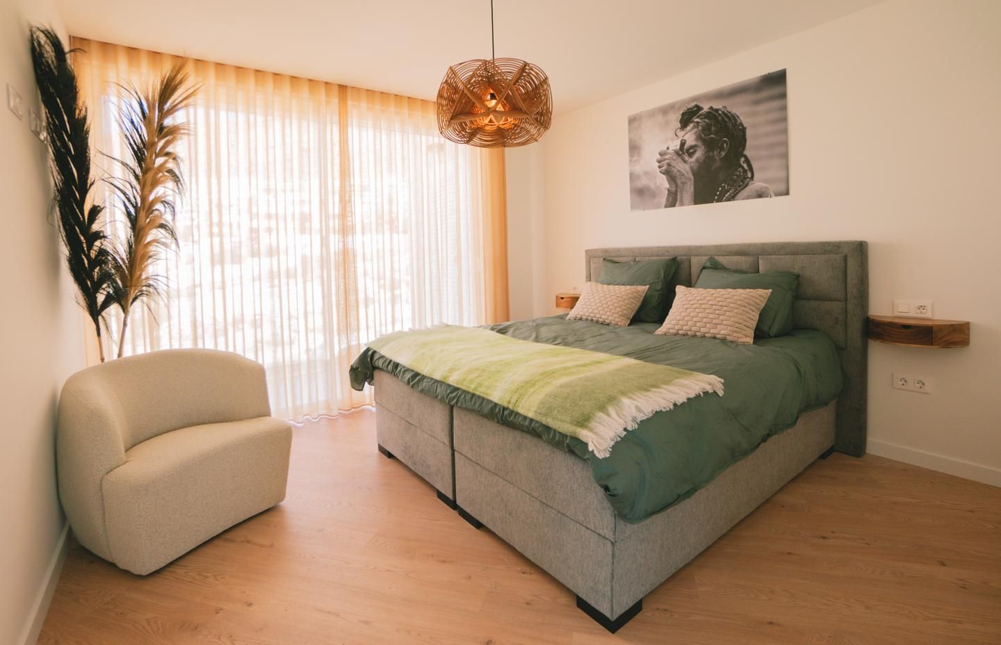 Bright bedroom with a large gray upholstered bed in green bedding, beige throw pillows, and a woven pendant light above. A cream rounded chair and tall plant sit by a sheer-curtained window, creating a relaxing space.