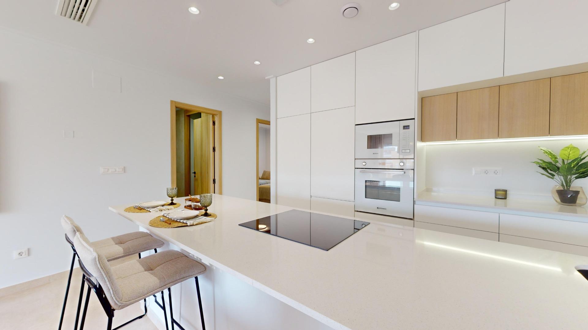 Modern white kitchen with a large island, built‑in microwave and oven, and white cabinetry; wooden wall cabinets above a light backsplash; plant and small candle on the counter to the right.