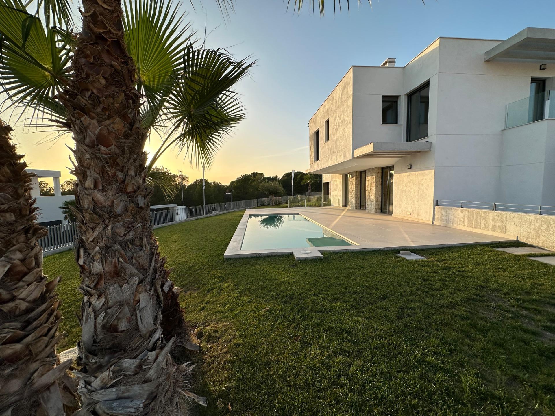 Modern white villa with a rectangular pool, palm trees, and a green lawn at sunset.