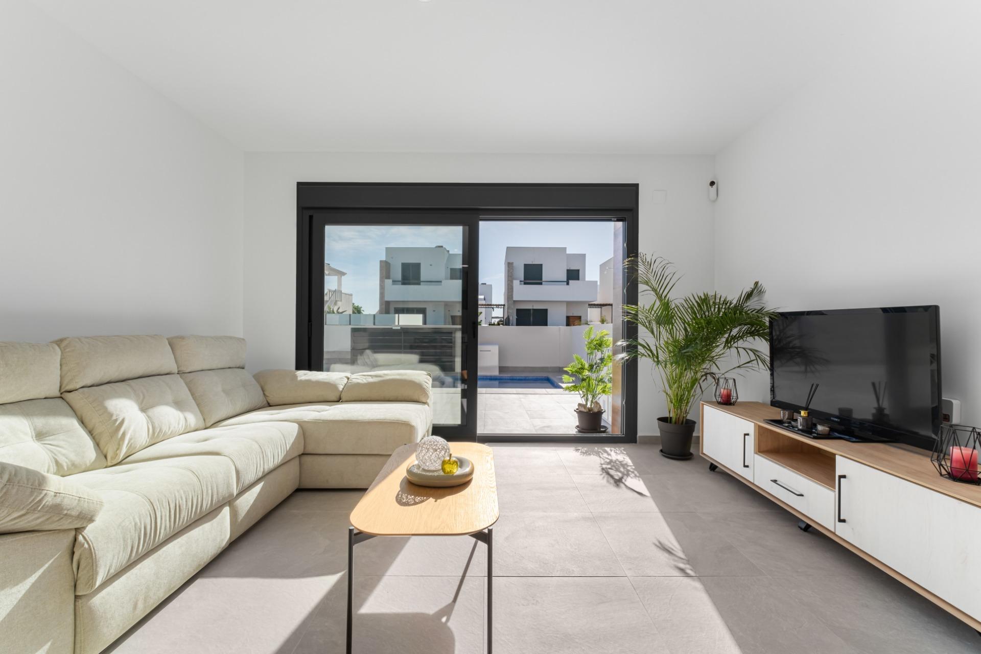 Bright living room with a beige sectional sofa, wooden coffee table, flat-screen TV, and several potted plants; large sliding glass doors reveal a patio and pool outdoors while natural light fills the space.