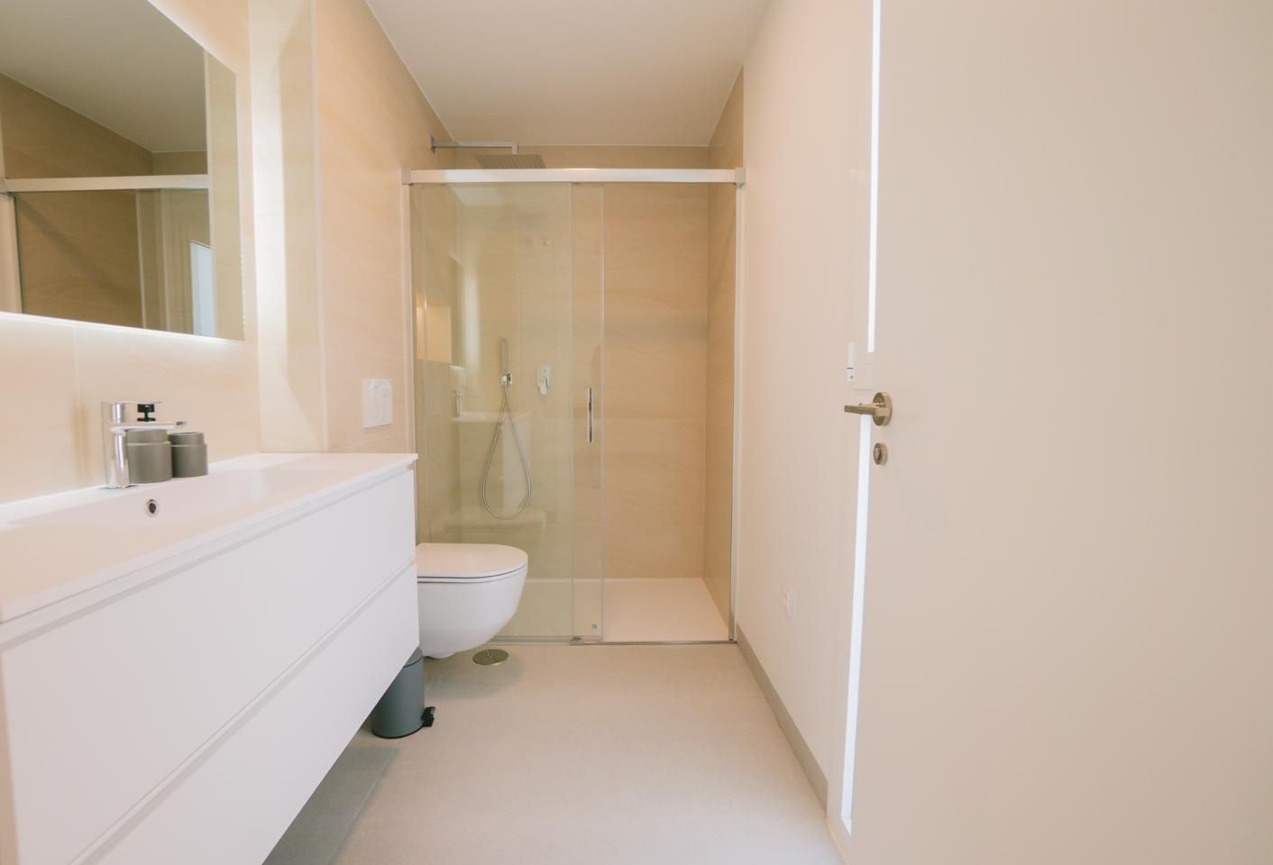 Bright modern bathroom with a white vanity, faucet, and mirror; glass shower enclosure at the back and a wall-hung toilet between them