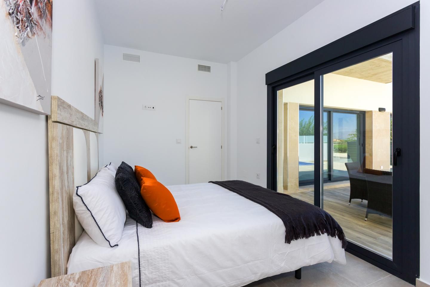 Modern bedroom with white bedding, orange and black throw pillows, and a large black-framed sliding glass door to an outdoor patio area.