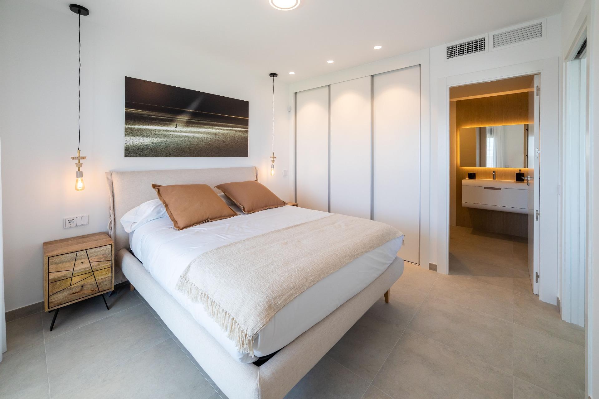 Contemporary bedroom with a beige upholstered bed, tan pillows, wooden nightstand, and hanging pendant lights; white sliding wardrobe doors across the wall.