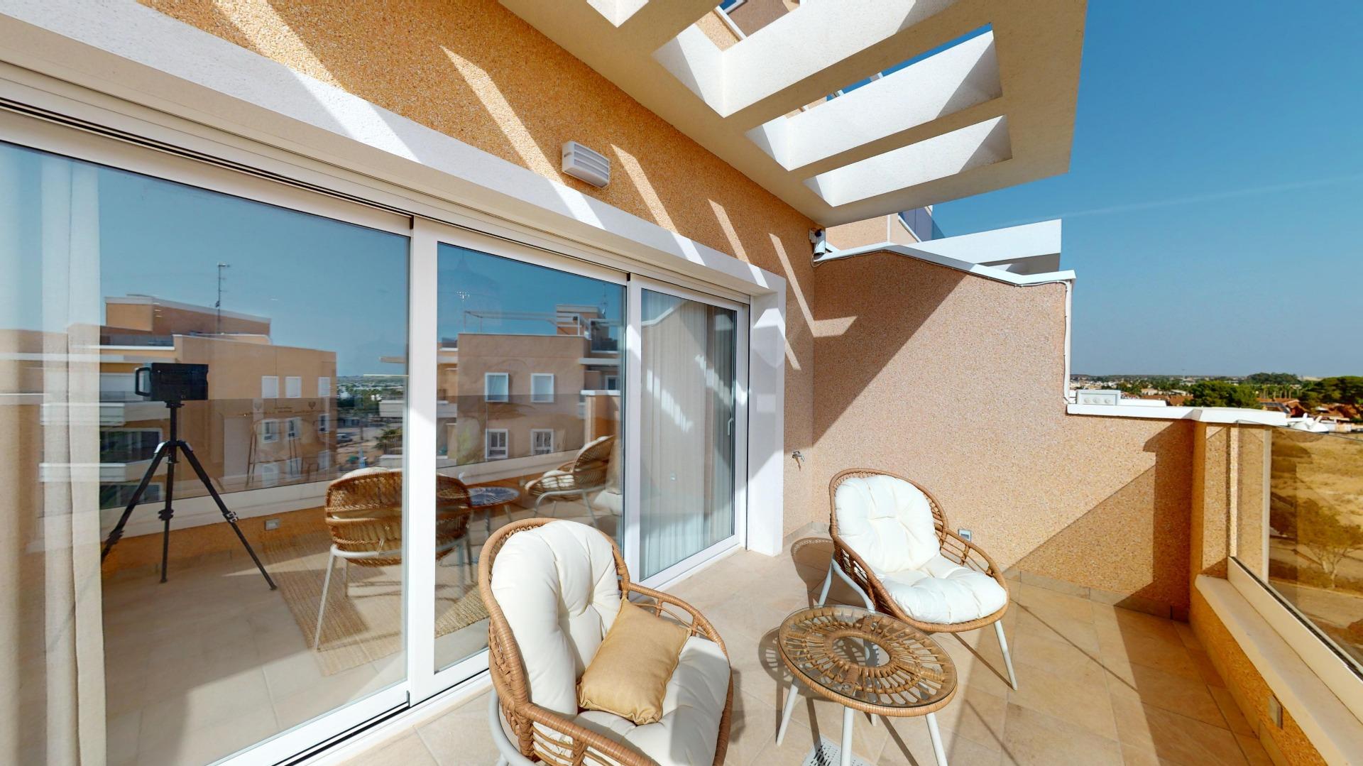 Sunlit apartment balcony with cushioned wicker chairs and a glass-top table, adjacent sliding glass doors reflecting nearby buildings and blue sky.