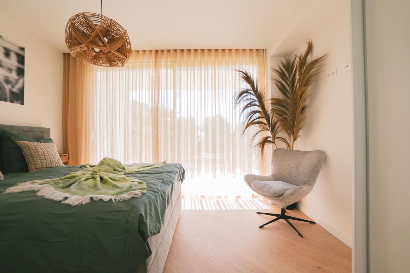 Sunlit bedroom with a green bedspread, wicker pendant lamp, and a gray swivel chair by a large window with sheer curtains.