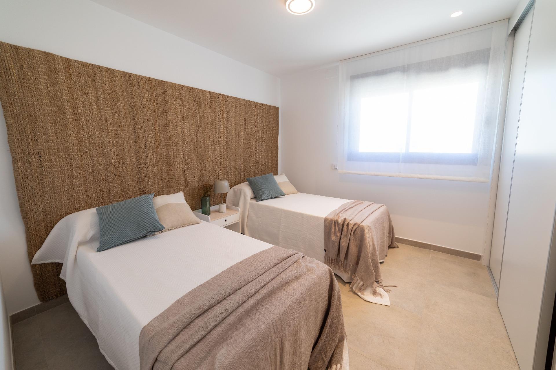 Twin bedroom with a large woven headboard between two single beds, white bedding, beige throws, and a shared nightstand with a lamp; a bright window with sheer curtains to the right.
