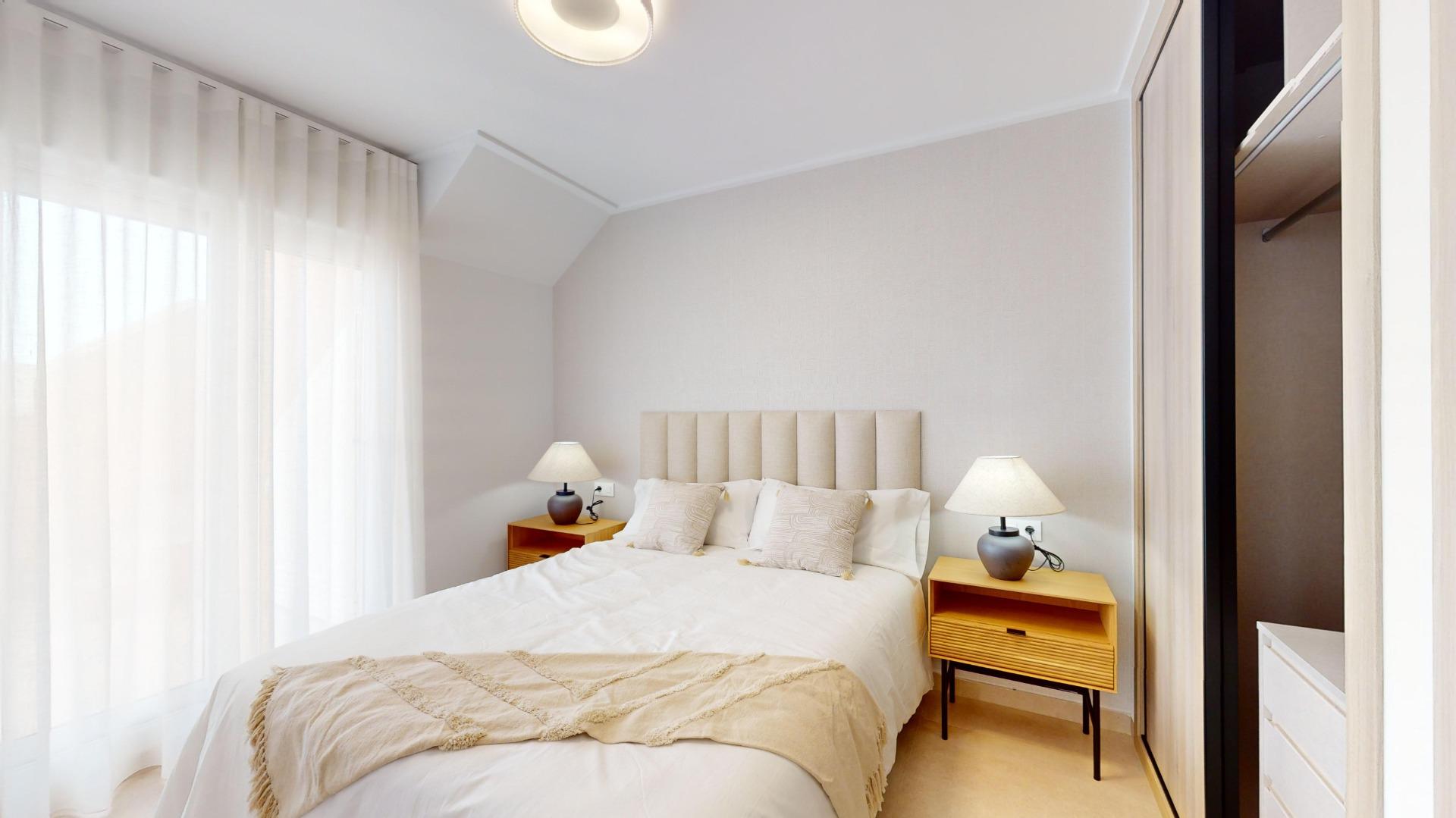 Bright bedroom with a large bed featuring a beige upholstered headboard, two wooden nightstands with lamps, and sheer white curtains along a wall of windows on the left.