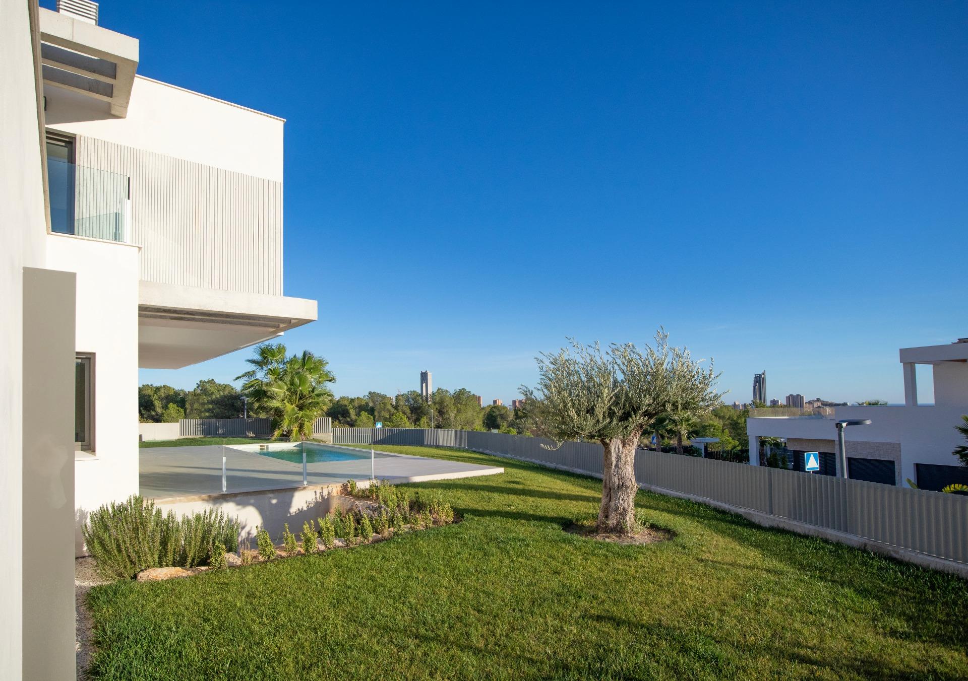 Modern white apartment building with a glass balcony, pool deck, and green lawn under a clear blue sky.