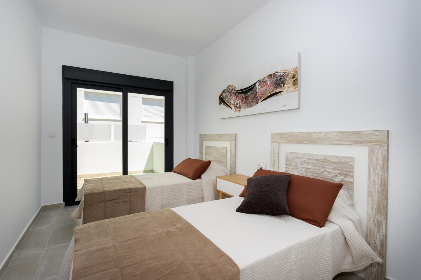 Twin bedroom with two single beds, beige bedding, and rustic headboards in a bright room with a black-framed glass door to a balcony.